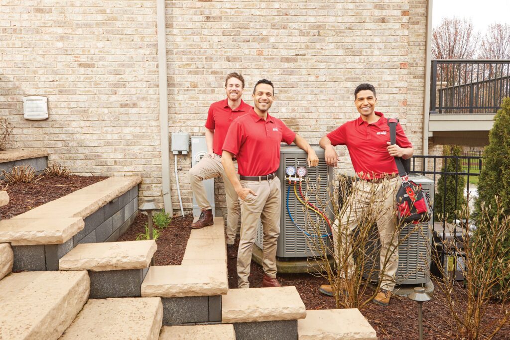 Three HVAC technicians in red polos leaning against an outdoor air conditioner